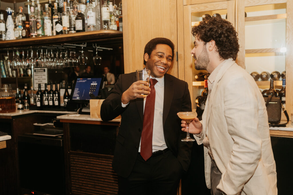 Two men lean by the bar with cocktails laughing together at a wedding reception. 