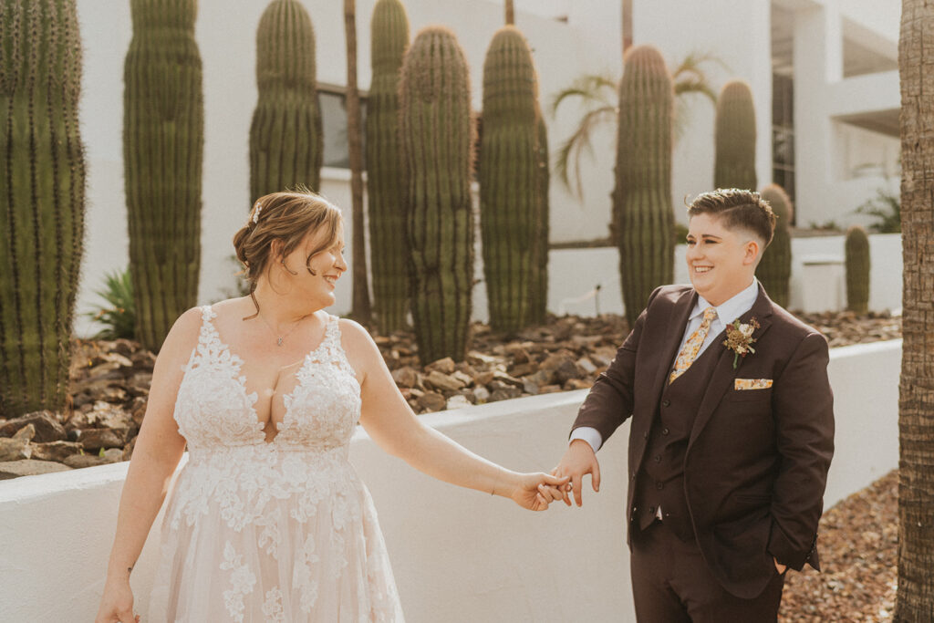 Two brides holding hands in front of white buildings and cacti wearing a dress and suit. 