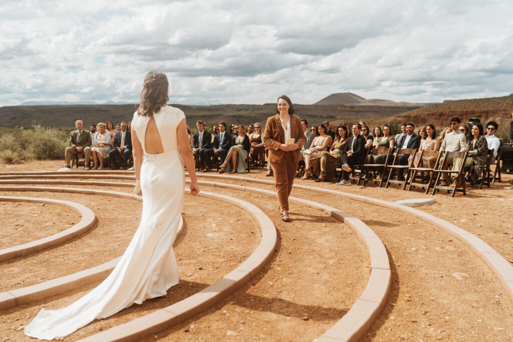 Two brides walk through a labyrinth to get to their wedding ceremony with views of the red rocks of Zion nation park behind them. 