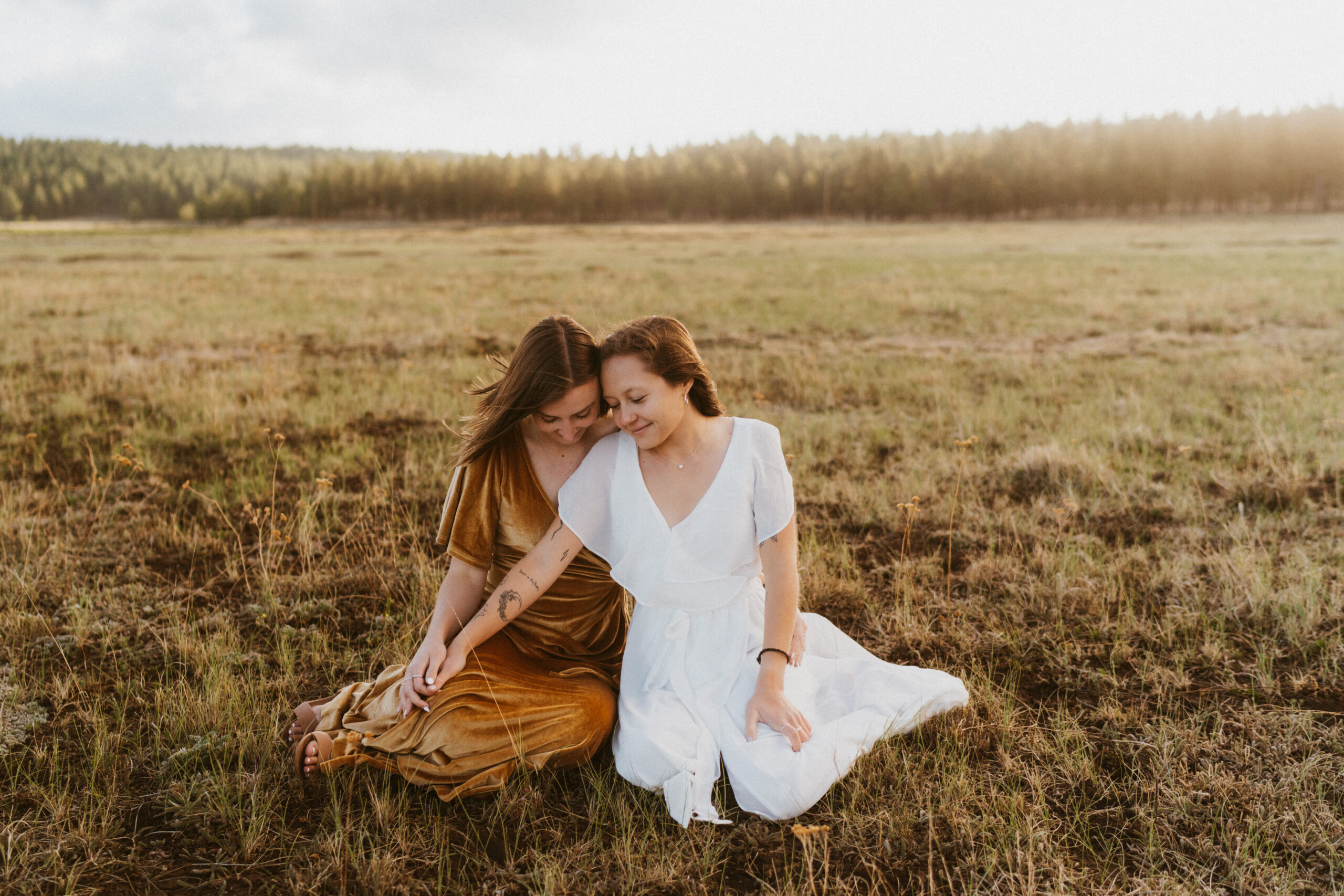 Two women, one in a white dress and the other in a mustard dress sit in a field holding hands