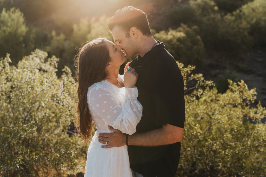 A woman in a white dress leans into kiss a man in a black shirt in the mountains 