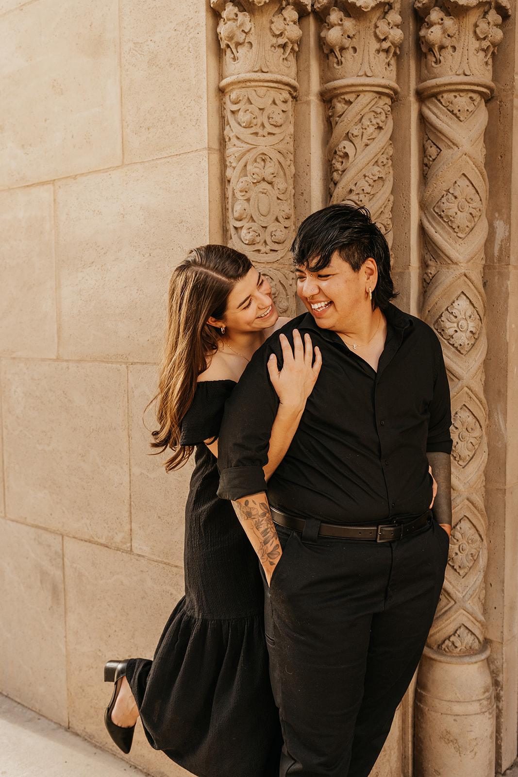 Two partners embrace by the wall of a building, both are wearing black clothing