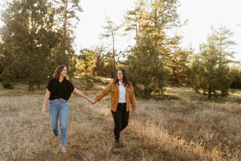 Two women holding hands walking in a field with pine trees behind them
