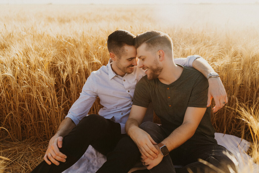Two men sitting in a wheat field, one has his arm around the other 