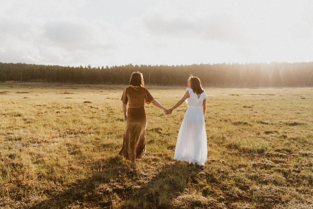 Two women in dresses walk through a field, surrounded by pine trees. 