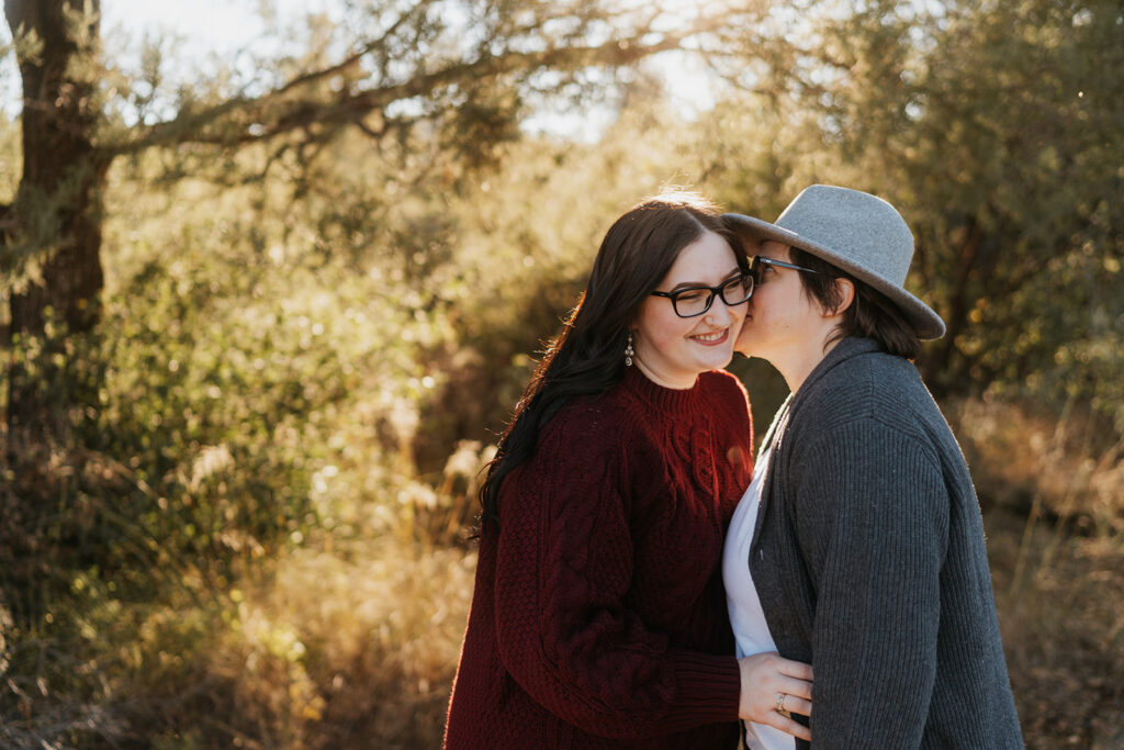 Two women embrace and one kisses the other on the cheek as they stand amongst pine trees and bushes. 