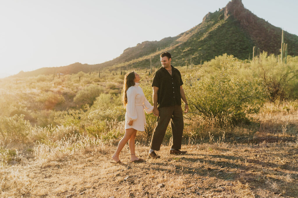 A woman and a man walk along a filed with a tall mountain and cacti behind them.
