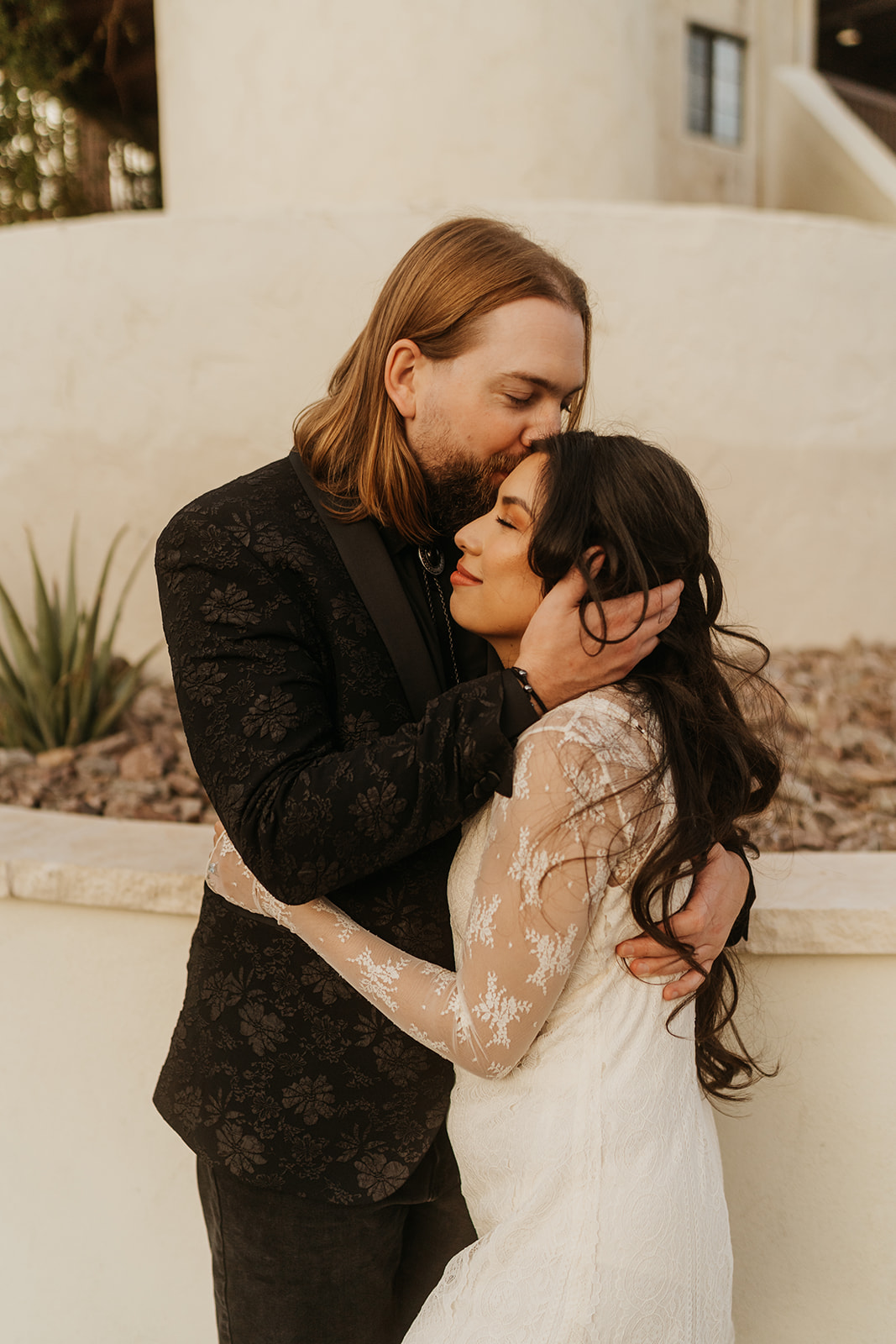 A tall man with red shoulder length hair and a shorter woman with long dark hair embrace, the man kisses the woman on the head.