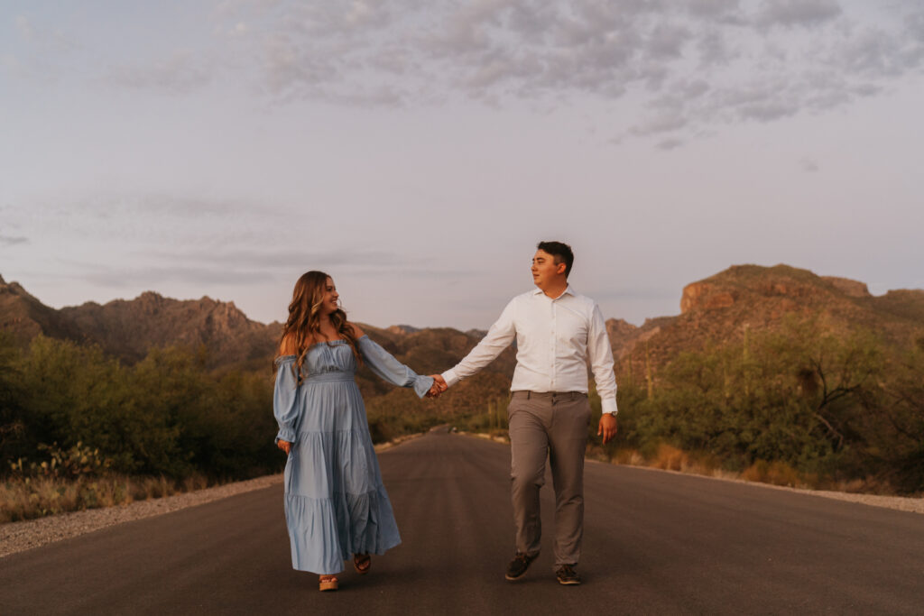 A man and a woman walk toward the camera, holding hands. They are framed with mountains in the background at dusk. 