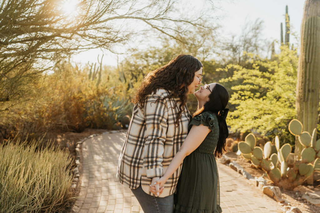 Two woman hold hands and lean in for a kiss surrounded by desert greenery.