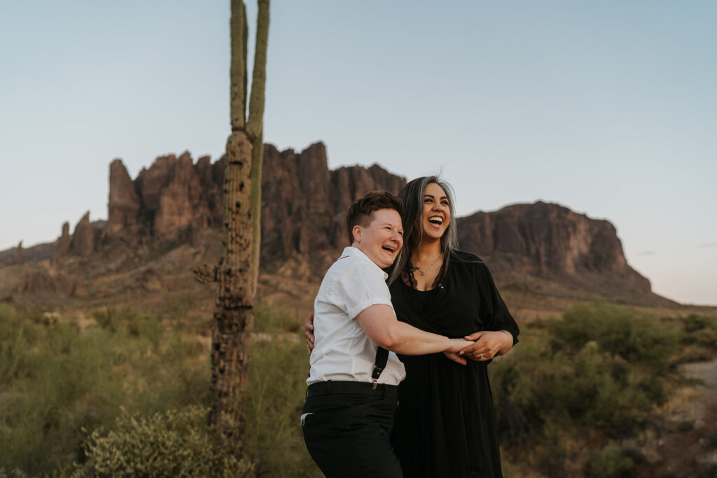 Two woman standing in from of the Superstition Mountains hold each other and laugh while looking in the distance.