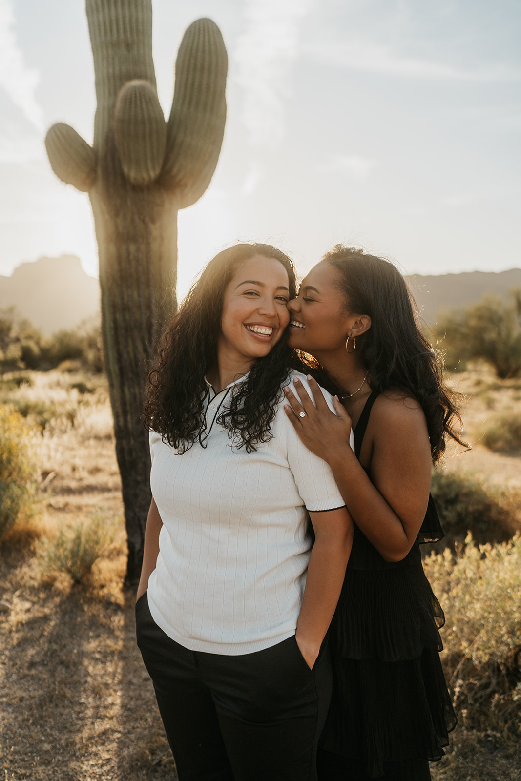 Two women standing in the desert with a cactus behind them. One is kissing the cheek of the other.